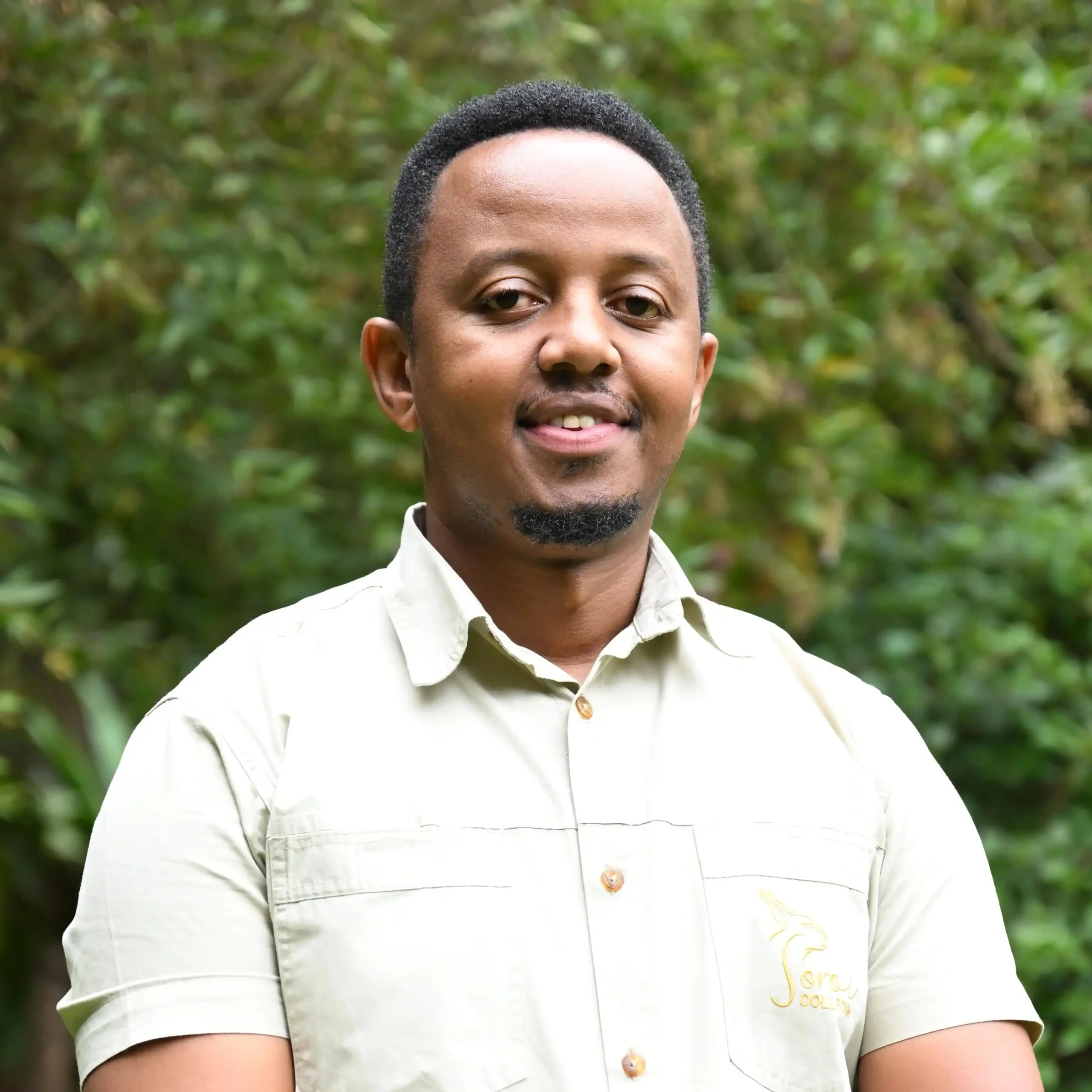 Close-up of a man outdoors in a light button-up shirt with green foliage in the background.