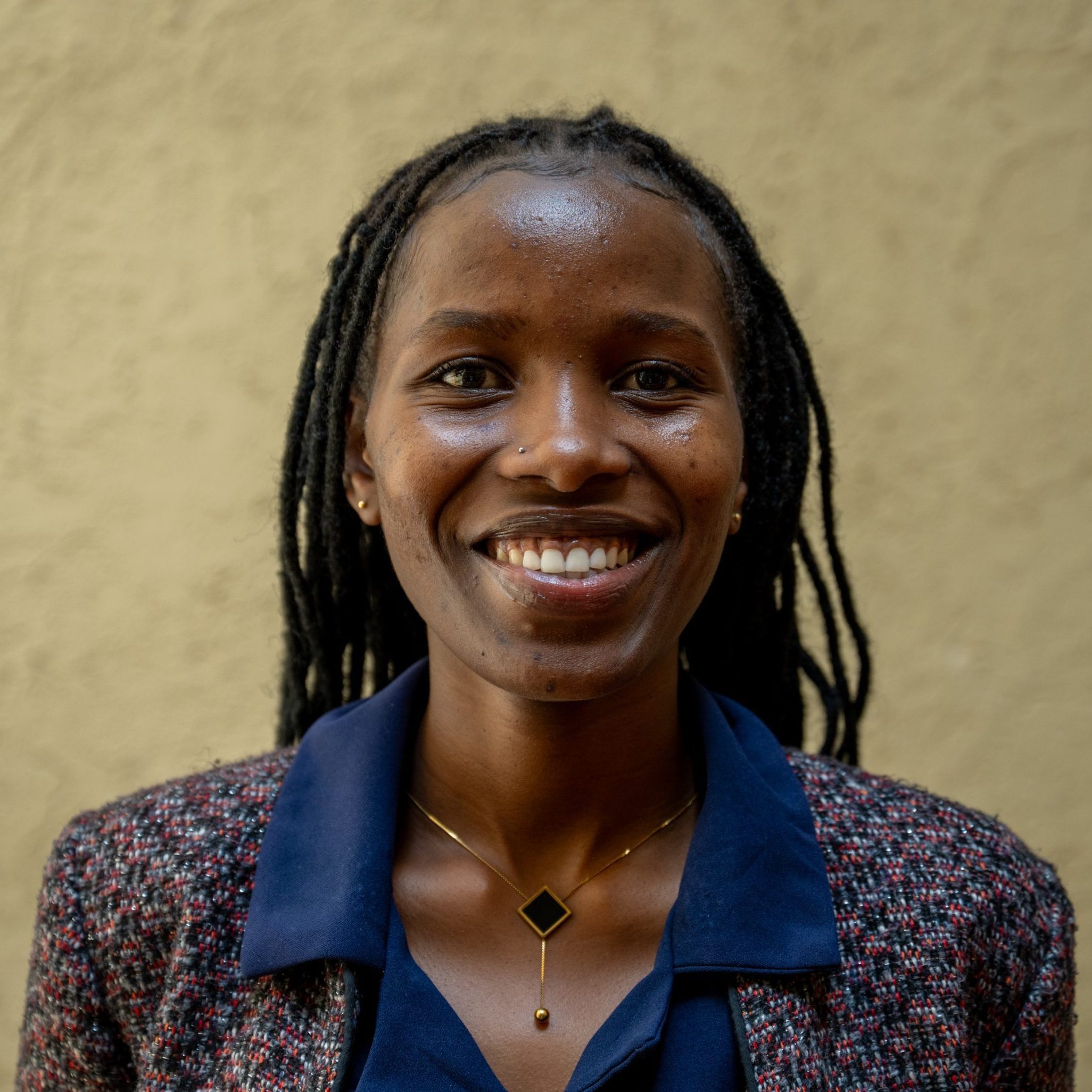 Close-up of a smiling woman with braided hair, wearing a navy scarf and a textured jacket against a beige wall.