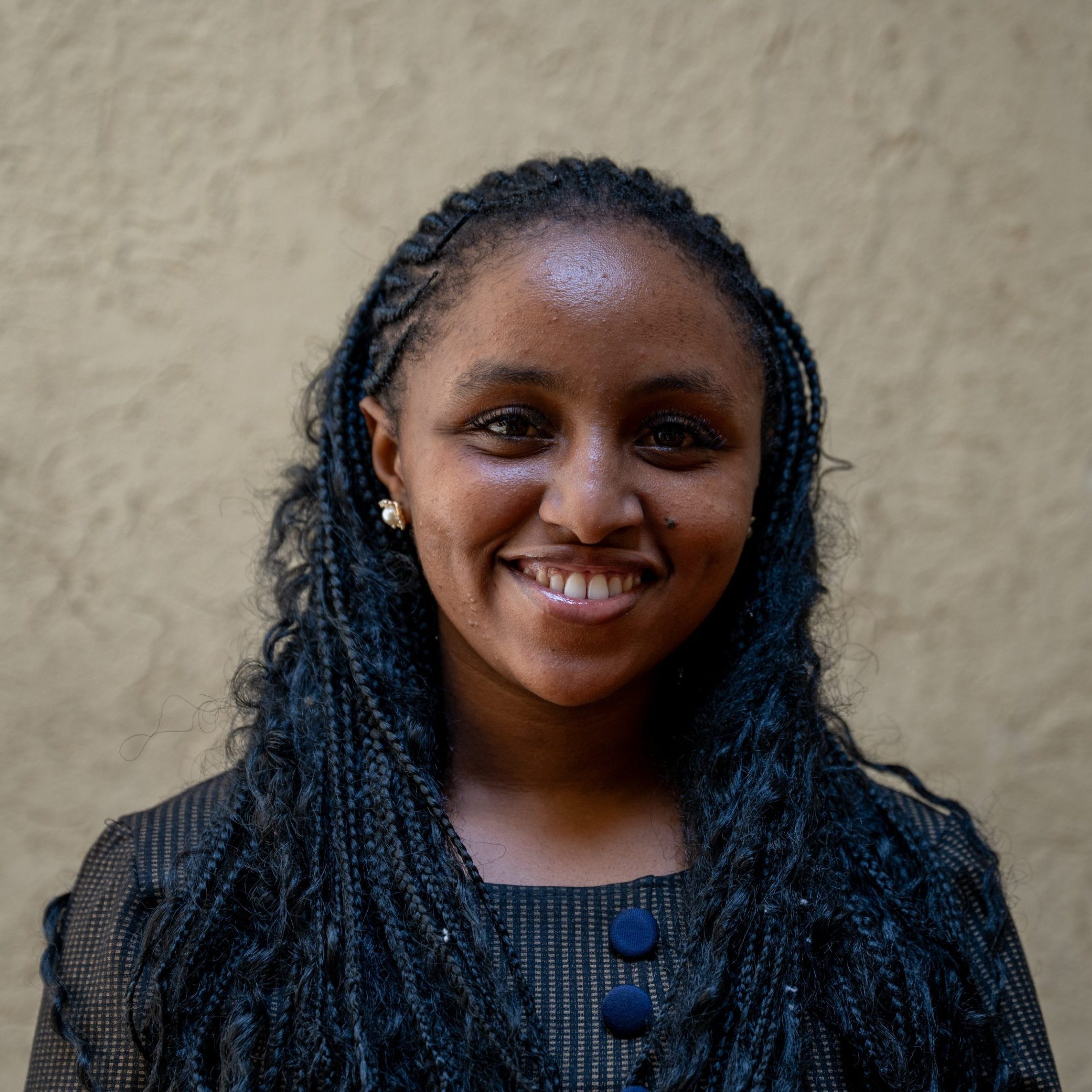 Smiling woman with braided hair against a beige wall, wearing a dark striped top with blue buttons.