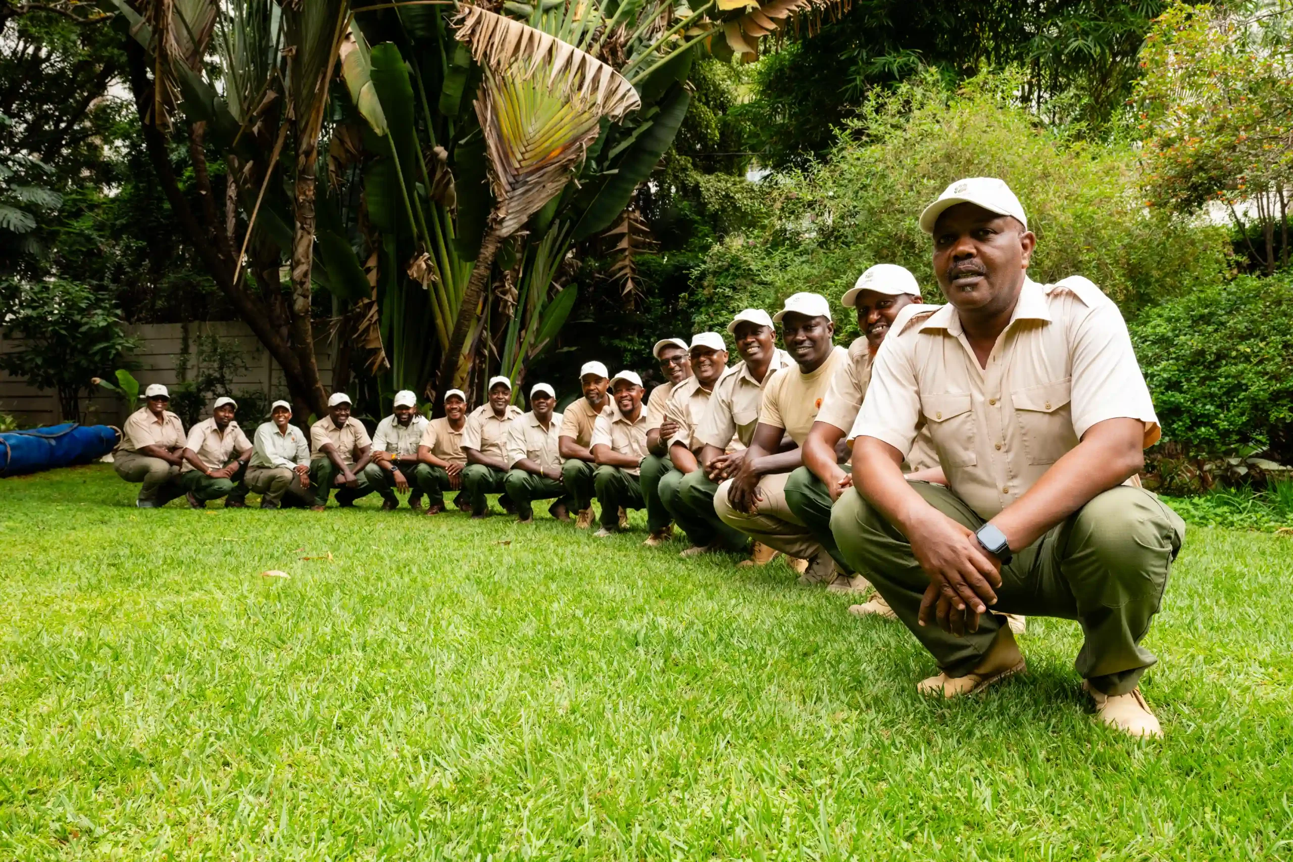 Group of uniformed men squatting in a grassy garden, posing for a photo outdoors.