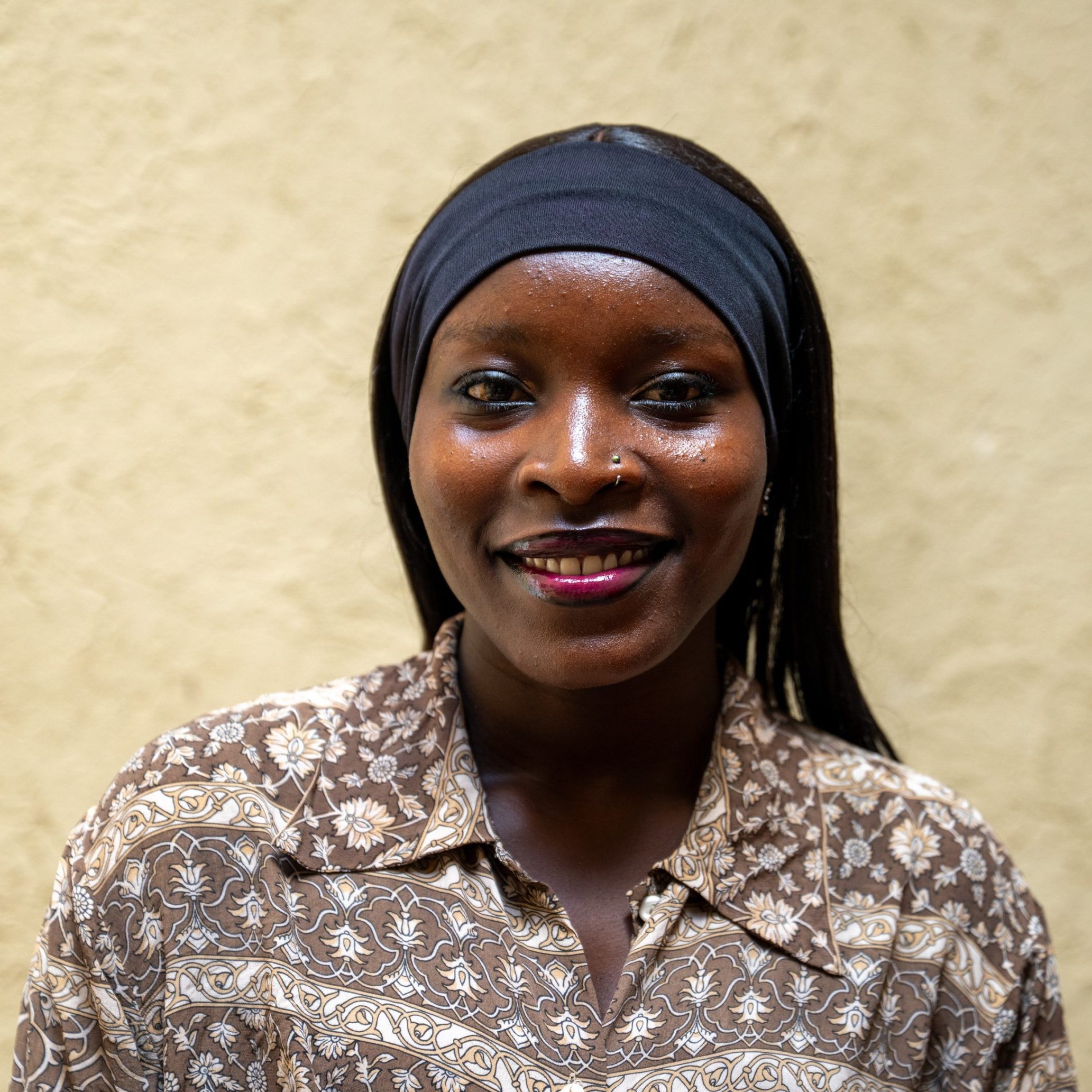 Close-up portrait of a smiling woman wearing a dark headwrap and a brown patterned blouse against a beige wall.