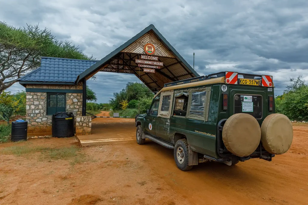 Entrance gate to Lumo Community Wildlife Conservancy; a green safari vehicle parked on a dirt path beneath a cloudy sky.