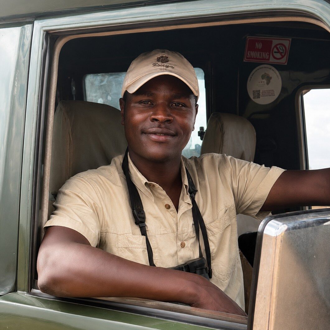 Man in a beige shirt and cap sits in the driver’s seat of a green vehicle, facing the camera.