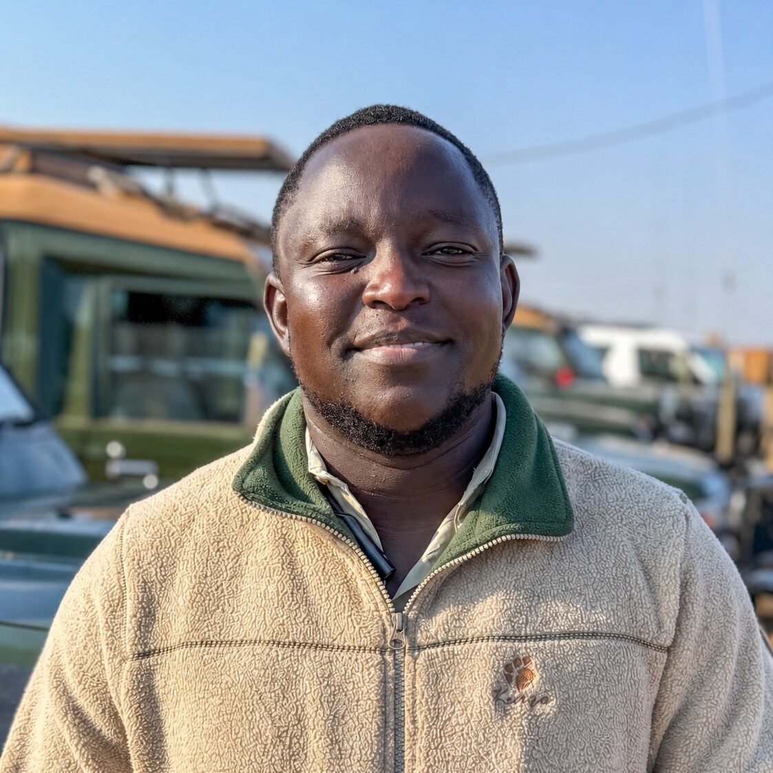 Smiling man outdoors wearing a beige fleece jacket with a green collar, with cars in the background under a clear blue sky