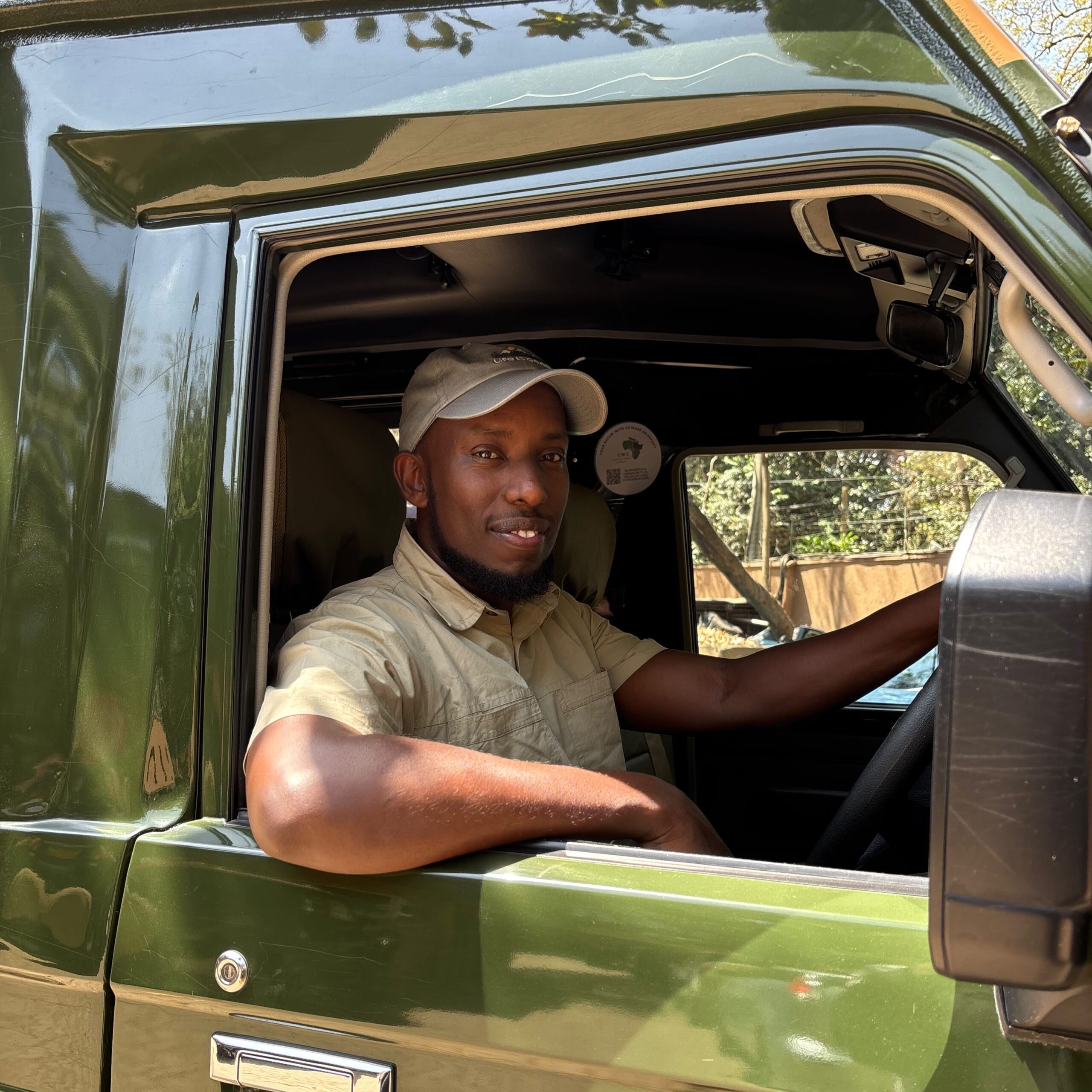 Man wearing a beige shirt and cap sits in the driver’s seat of a green pickup with the window down, looking at the camera.